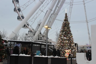 A large decorated Christmas tree adorned with lights and ornaments is set in an outdoor area near a ferris wheel. Snow covers parts of the scene, creating a wintery atmosphere. There are people gathered around, wrapped in winter clothing, suggesting a festive event. Nearby structures and lamp posts are also visible.