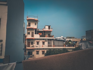 A multi-story residential building with balconies and an orange accent on the edges stands prominently. The structure is surrounded by other buildings, indicating a densely populated urban area. Laundry is visible hanging on one of the balconies, and the sky is clear with a hint of aeriness.
