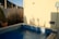 A small outdoor pool surrounded by beige walls and tall grass. There is a blue pool net leaning against the wall, and a sign indicating the pool depth along with safety instructions. The water in the pool is calm, reflecting the sunlight.