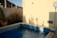 A small outdoor pool surrounded by beige walls and tall grass. There is a blue pool net leaning against the wall, and a sign indicating the pool depth along with safety instructions. The water in the pool is calm, reflecting the sunlight.