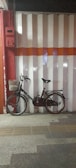 A vintage bicycle is leaning against a garage door with vertical pink and white stripes and a horizontal orange stripe across the middle. The bicycle has a metal basket attached to the front and a rear rack, with noticeable wear and slight rust, highlighting its use and age. The ground is textured with tactile paving in front of the bicycle.