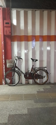 A vintage bicycle is leaning against a garage door with vertical pink and white stripes and a horizontal orange stripe across the middle. The bicycle has a metal basket attached to the front and a rear rack, with noticeable wear and slight rust, highlighting its use and age. The ground is textured with tactile paving in front of the bicycle.