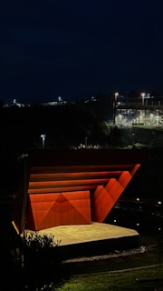 An outdoor stage with a geometric, angular design is illuminated at night. The structure is primarily composed of vibrant orange panels that stand out against the dark surroundings. The environment includes a grassy area and dimly lit trees, while distant buildings are faintly visible under artificial lights.
