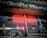 A storefront window with a bright red neon sign advertising books, CDs, videos, and comics. Inside the window display, there are various items including musical instruments like a guitar and a drum, along with vintage records. The exterior is somewhat retro, with a rustic sign on top and reflections from the street.