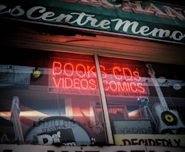 A storefront window with a bright red neon sign advertising books, CDs, videos, and comics. Inside the window display, there are various items including musical instruments like a guitar and a drum, along with vintage records. The exterior is somewhat retro, with a rustic sign on top and reflections from the street.