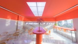 A modern, brightly lit interior with vibrant orange and white color scheme features a central counter with a decorative plant. Transparent chairs line a bar against large windows showing a partial outdoor view.