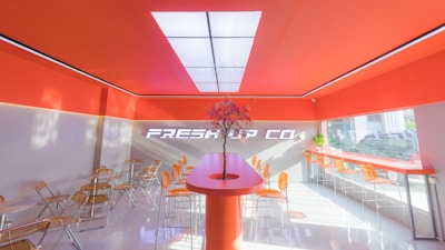 A modern, brightly lit interior with vibrant orange and white color scheme features a central counter with a decorative plant. Transparent chairs line a bar against large windows showing a partial outdoor view.