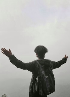 A person with arms outstretched stands at the edge of a hazy landscape, suggesting a sense of freedom or triumph. The foggy background creates a serene and contemplative atmosphere.