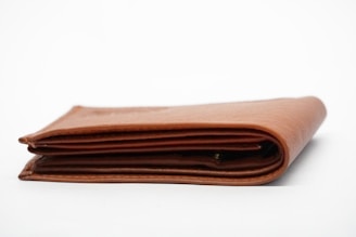 A brown leather wallet is folded and placed on a plain white background. The wallet's edges and stitching are visible, indicating simple and classic craftsmanship.