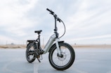 A sleek, modern electric bike stands on a paved surface with a vast, open background. The bike has a sturdy frame, large tires, and is equipped with a suspension fork. The brand name is visible on the frame, and the bike seems ready for both urban and off-road adventures.