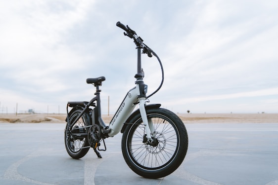 A sleek, modern electric bike stands on a paved surface with a vast, open background. The bike has a sturdy frame, large tires, and is equipped with a suspension fork. The brand name is visible on the frame, and the bike seems ready for both urban and off-road adventures.