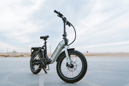 A sleek, modern electric bike stands on a paved surface with a vast, open background. The bike has a sturdy frame, large tires, and is equipped with a suspension fork. The brand name is visible on the frame, and the bike seems ready for both urban and off-road adventures.