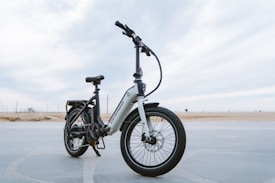 A sleek, modern electric bike stands on a paved surface with a vast, open background. The bike has a sturdy frame, large tires, and is equipped with a suspension fork. The brand name is visible on the frame, and the bike seems ready for both urban and off-road adventures.