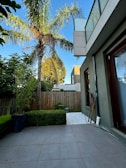 A backyard area with a tiled patio, surrounded by well-maintained greenery including a large palm tree and trimmed hedges. A tall wooden fence separates the yard from neighboring properties. The exterior of a modern building with large windows is visible on the right. The sky is clear and blue.
