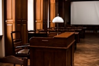 A dimly lit room with wooden furniture, including a desk and chairs. A white lamp sits atop the desk, and there's a large blank screen in the background. The flooring is a parquet pattern, and there are tall wooden cabinets along the wall.