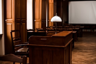 A dimly lit room with wooden furniture, including a desk and chairs. A white lamp sits atop the desk, and there's a large blank screen in the background. The flooring is a parquet pattern, and there are tall wooden cabinets along the wall.