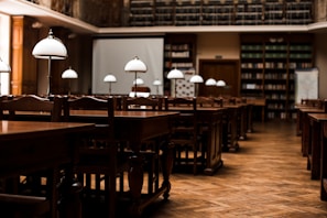 A quiet, empty library with wooden tables and chairs arranged in neat rows. Elegant white lamps with metal stands are placed on each table, casting a warm glow. Bookshelves lining the walls are filled with books, and a large projection screen is set up at the front of the room.