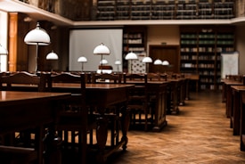 A quiet, empty library with wooden tables and chairs arranged in neat rows. Elegant white lamps with metal stands are placed on each table, casting a warm glow. Bookshelves lining the walls are filled with books, and a large projection screen is set up at the front of the room.