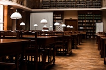 A quiet, empty library with wooden tables and chairs arranged in neat rows. Elegant white lamps with metal stands are placed on each table, casting a warm glow. Bookshelves lining the walls are filled with books, and a large projection screen is set up at the front of the room.