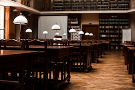 A quiet, empty library with wooden tables and chairs arranged in neat rows. Elegant white lamps with metal stands are placed on each table, casting a warm glow. Bookshelves lining the walls are filled with books, and a large projection screen is set up at the front of the room.