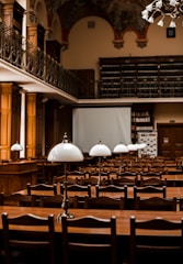 A large, elegant library or reading room featuring rows of wooden tables and chairs. Each table is equipped with classic white lamps. The room has high ceilings with ornate decorations and a balcony lined with bookshelves filled with numerous books. The atmosphere is scholarly and inviting.