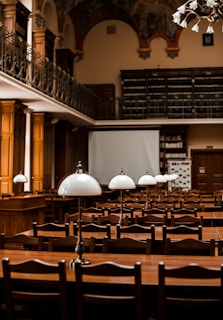 A large, elegant library or reading room featuring rows of wooden tables and chairs. Each table is equipped with classic white lamps. The room has high ceilings with ornate decorations and a balcony lined with bookshelves filled with numerous books. The atmosphere is scholarly and inviting.