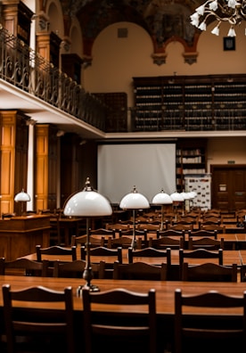 A large, elegant library or reading room featuring rows of wooden tables and chairs. Each table is equipped with classic white lamps. The room has high ceilings with ornate decorations and a balcony lined with bookshelves filled with numerous books. The atmosphere is scholarly and inviting.