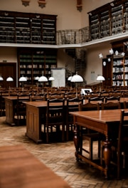 Rows of wooden tables and chairs fill a spacious, historic library with high ceilings. The walls are lined with tall bookshelves filled with books. Delicate light fixtures hang over the tables, and a spiral staircase leads to an upper level with a balcony and more bookshelves.