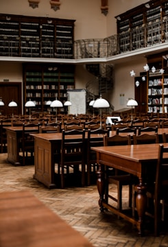 Rows of wooden tables and chairs fill a spacious, historic library with high ceilings. The walls are lined with tall bookshelves filled with books. Delicate light fixtures hang over the tables, and a spiral staircase leads to an upper level with a balcony and more bookshelves.