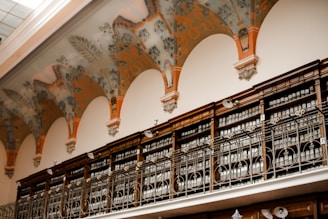 A high-ceilinged room with ornate decorative arches featuring intricate patterns and flourishes. Below the arches, there is a series of wooden shelves with ornately designed metal railings. The shelves are lined with books or files, organized in a neat and orderly manner.
