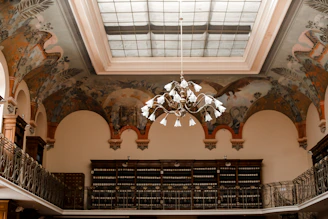 An ornate ceiling with a large skylight is adorned with detailed murals depicting natural and historical scenes. Below, a two-tiered library is lined with shelves filled with books, featuring intricate wrought iron balconies and railings. A grand chandelier with numerous white lampshades hangs from the ceiling, adding an elegant touch to the space.