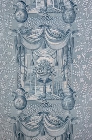 A detailed and intricate wallpaper pattern with symmetrical design elements. Two large urns feature decorative patterns and are flanked by stylized foliage. A central vase holds an ornate bouquet of flowers, framed by draped fabric and tassels. The background displays lattice patterns, and scenes reminiscent of classical art appear within the frames.