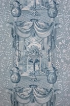 A detailed and intricate wallpaper pattern with symmetrical design elements. Two large urns feature decorative patterns and are flanked by stylized foliage. A central vase holds an ornate bouquet of flowers, framed by draped fabric and tassels. The background displays lattice patterns, and scenes reminiscent of classical art appear within the frames.
