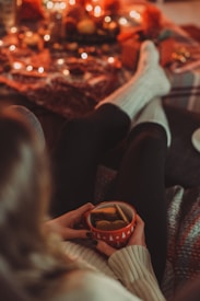 A person wearing cozy socks is sitting comfortably with a mug filled with tea and biscuits, creating a warm and inviting atmosphere. The background is decorated with soft blankets and illuminated by soft, warm string lights, contributing to a festive and snug setting.