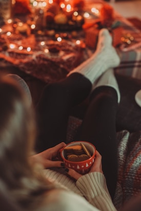 A person wearing cozy socks is sitting comfortably with a mug filled with tea and biscuits, creating a warm and inviting atmosphere. The background is decorated with soft blankets and illuminated by soft, warm string lights, contributing to a festive and snug setting.