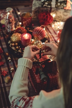 A cozy, festive holiday scene featuring a person wrapping a gift with a red wrapping paper decorated with small hearts. The setting includes a table covered with a plaid cloth adorned with Christmas ornaments like baubles, a lit candle, and seasonal decorations like pinecones and fairy lights. Various wrapped gifts and festive items add to the warm, ambient atmosphere.