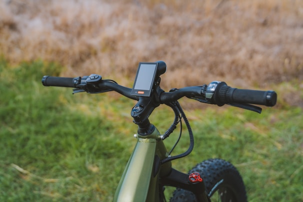 A close-up view of the handlebars and control panel of a modern electric bike, set against a blurred natural background with grass and earthy tones. The handlebars feature grip handles, a brake lever, and a display screen.