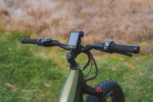 A close-up view of the handlebars and control panel of a modern electric bike, set against a blurred natural background with grass and earthy tones. The handlebars feature grip handles, a brake lever, and a display screen.
