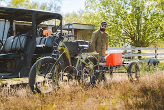 A man in outdoor clothing stands next to an electric bicycle equipped with a trailer carrying an orange cooler. Nearby, there is an off-road vehicle with several seats. The setting appears to be rural or countryside, with green trees, grass, and a fence visible in the background under bright daylight.