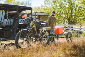 A man in outdoor clothing stands next to an electric bicycle equipped with a trailer carrying an orange cooler. Nearby, there is an off-road vehicle with several seats. The setting appears to be rural or countryside, with green trees, grass, and a fence visible in the background under bright daylight.