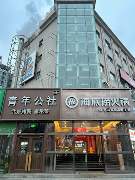 A multi-story building with a modern glass facade featuring a restaurant named Hai Di Lao Hot Pot on the ground floor. The building has a sleek design with prominent signage in both Chinese characters and an English logo. One side of the building features a tall, transparent glass section, likely housing an elevator shaft.