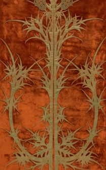 An intricate pattern featuring symmetrical, stylized floral or thistle motifs in a muted green color, set against a rich, rust-colored background. The design is complex and detailed, suggesting an artistic or decorative textile.