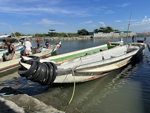A group of small, motorized fishing boats is docked by a calm, narrow river. Five individuals are seen interacting on or near the boats; they appear to be engaged in a task involving the boats, possibly maintenance or preparation for a trip. The setting includes a backdrop of greenery and a clear blue sky, with the reflection of the boats shimmering in the water. A coiled bundle of black tubing or ropes lies in one of the boats.