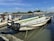A group of small, motorized fishing boats is docked by a calm, narrow river. Five individuals are seen interacting on or near the boats; they appear to be engaged in a task involving the boats, possibly maintenance or preparation for a trip. The setting includes a backdrop of greenery and a clear blue sky, with the reflection of the boats shimmering in the water. A coiled bundle of black tubing or ropes lies in one of the boats.