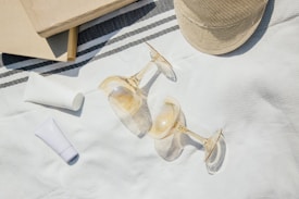 A neatly arranged outdoor scene featuring two books, two empty wine glasses, a straw hat, and two tubes of lotion or sunscreen, all placed on a textured white cloth with a striped pattern. The lighting suggests a sunny day, casting soft shadows and creating a relaxed, summery ambiance.
