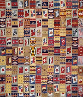 A vibrant patchwork quilt composed of various geometric patterns, flags, and symbolic designs. Each patch is unique and features a range of motifs including stripes, circles, and abstract shapes. The quilt is rich in color and symmetry, capturing a sense of cultural diversity and artistic craftsmanship.