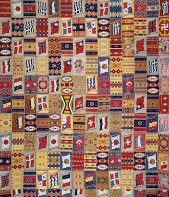 A vibrant patchwork quilt composed of various geometric patterns, flags, and symbolic designs. Each patch is unique and features a range of motifs including stripes, circles, and abstract shapes. The quilt is rich in color and symmetry, capturing a sense of cultural diversity and artistic craftsmanship.
