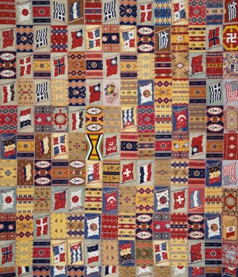 A vibrant patchwork quilt composed of various geometric patterns, flags, and symbolic designs. Each patch is unique and features a range of motifs including stripes, circles, and abstract shapes. The quilt is rich in color and symmetry, capturing a sense of cultural diversity and artistic craftsmanship.
