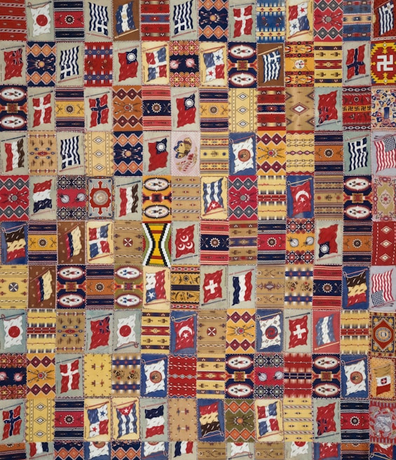 A vibrant patchwork quilt composed of various geometric patterns, flags, and symbolic designs. Each patch is unique and features a range of motifs including stripes, circles, and abstract shapes. The quilt is rich in color and symmetry, capturing a sense of cultural diversity and artistic craftsmanship.