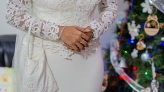 A person wearing an elegant white lace dress with intricate floral patterns, standing beside a decorated Christmas tree adorned with colorful lights and ornaments. The person's hands are adorned with rings and a bracelet.